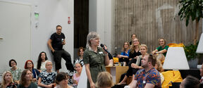A group of people sit and listen to a woman standing in the middle with a microphone in her hand.