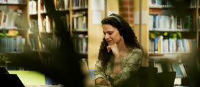 A person with curly hair is sitting in a library, wearing headphones and using a laptop. Bookshelves are in the background.